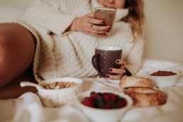 femme sur le lit qui regarde son téléphone devant son petit déjeuner avec des céréales et des fruits
