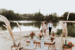 décoration de mariage d'automne au bord de l'eau. Décoration de pampa, table de mariage. Repas de mariage sur le sable. Fleurs de mariage, bouquet de mariage en pampa et roses orange. Robe de mariée bustier, fente sur la cuisse