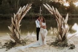 couple de mariés sous une arche de cérémonie en pampa. mariage en automne. robe de mariage bustier et traine. mariage au bord de l'eau